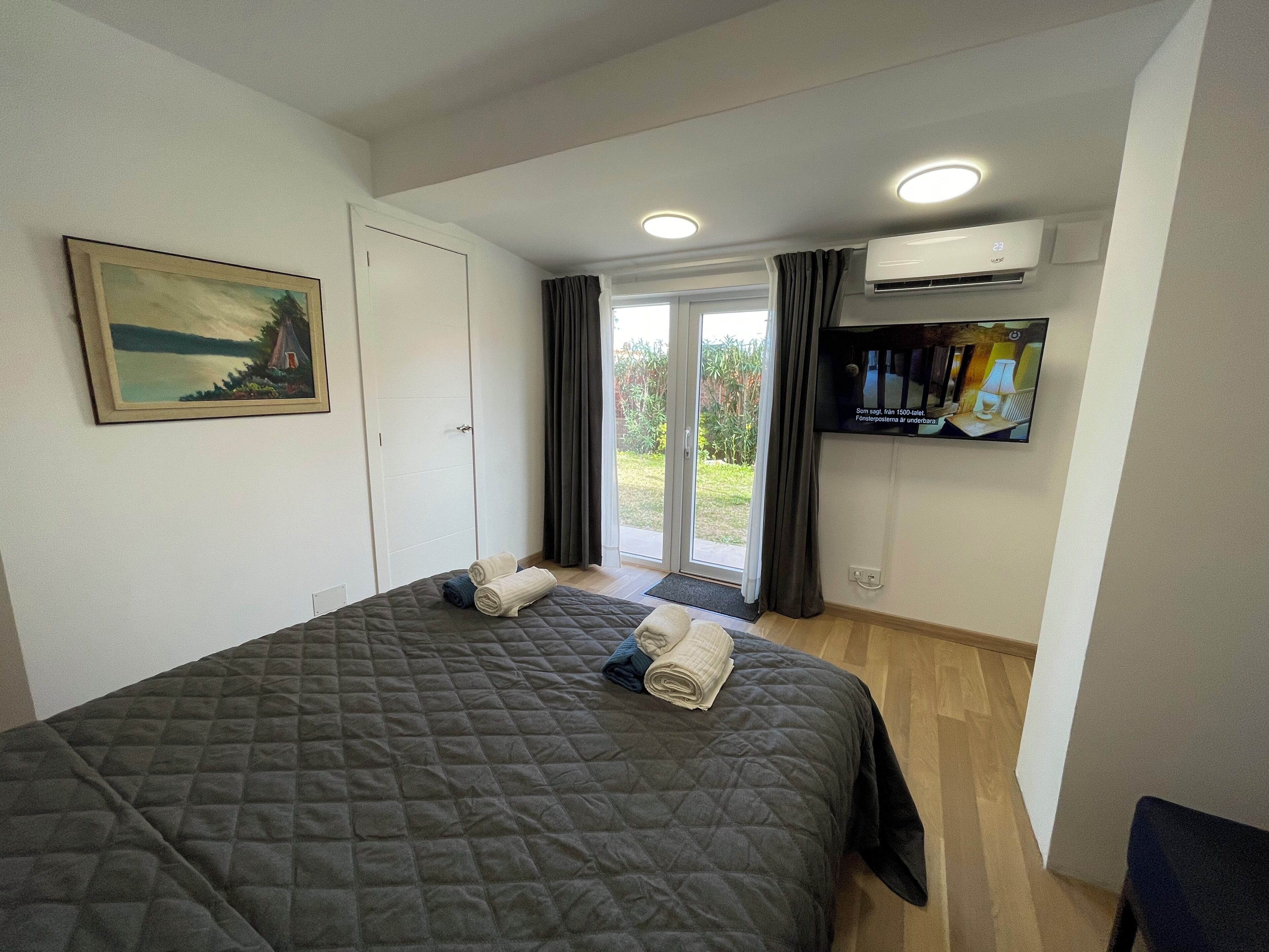A bedroom with a bed, a painting, an air conditioning unit, a television, and a sliding glass door to the outside.