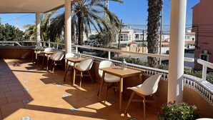 A covered outdoor seating area with tables and chairs, palm trees, and a view of buildings in the background. - Annas Garden (Torrevieja)