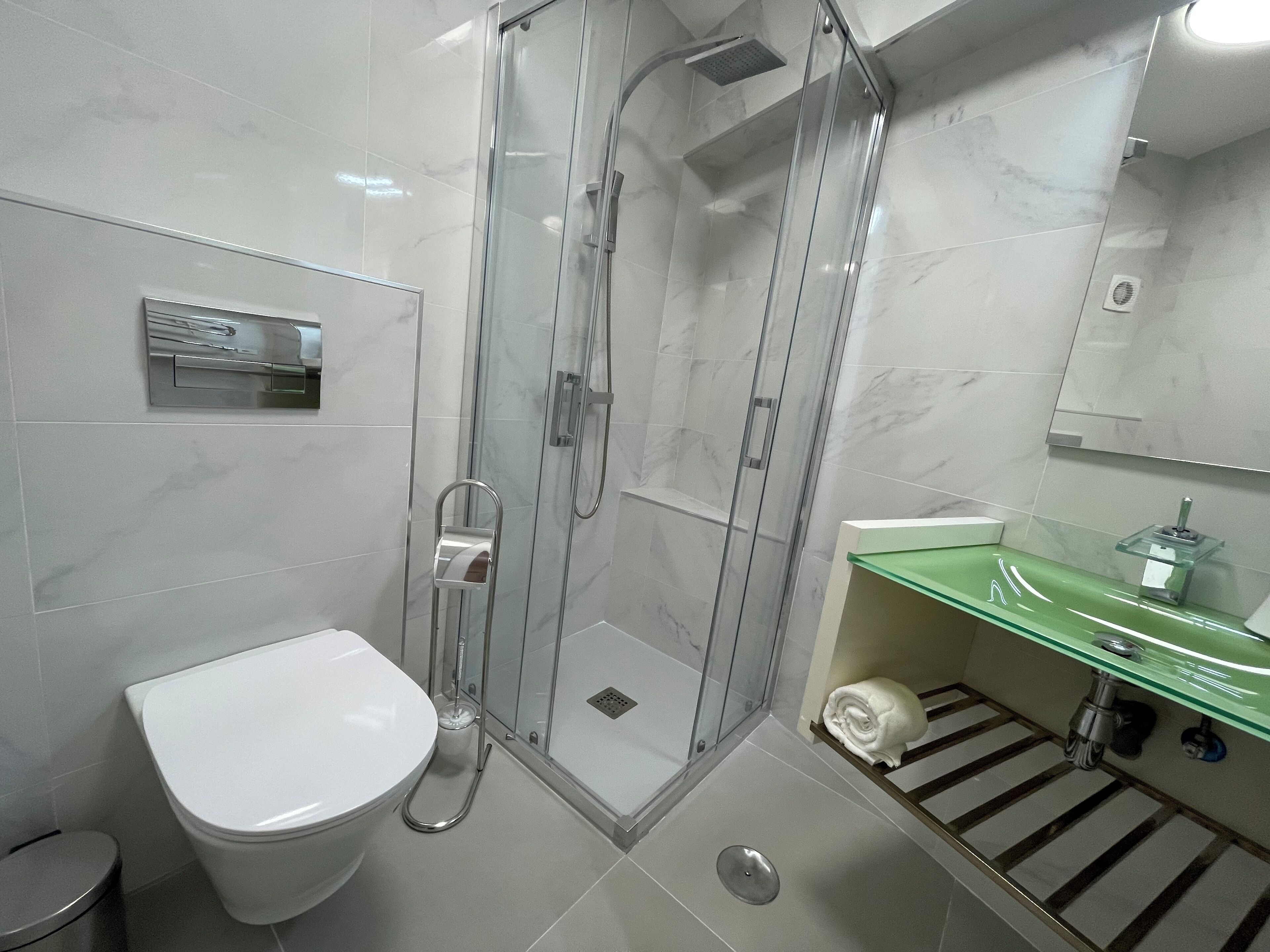 Een moderne badkamer met een douchecabine van glas, een wit toilet en een groene glazen wastafel met een spiegel erboven.