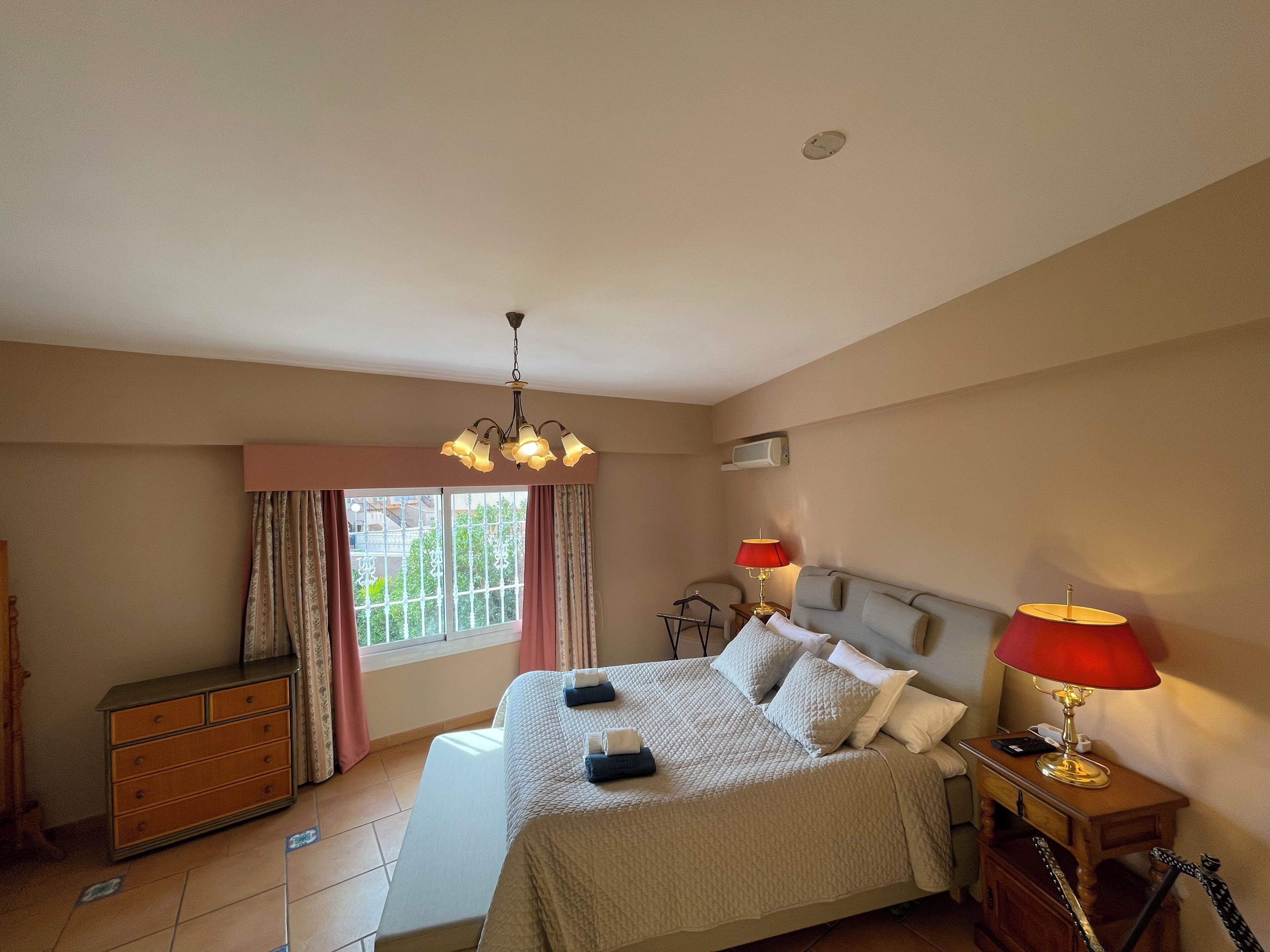 Een slaapkamer met een bed, nachtlampjes, een dressoir, een raam met gordijnen en een plafondlamp.
