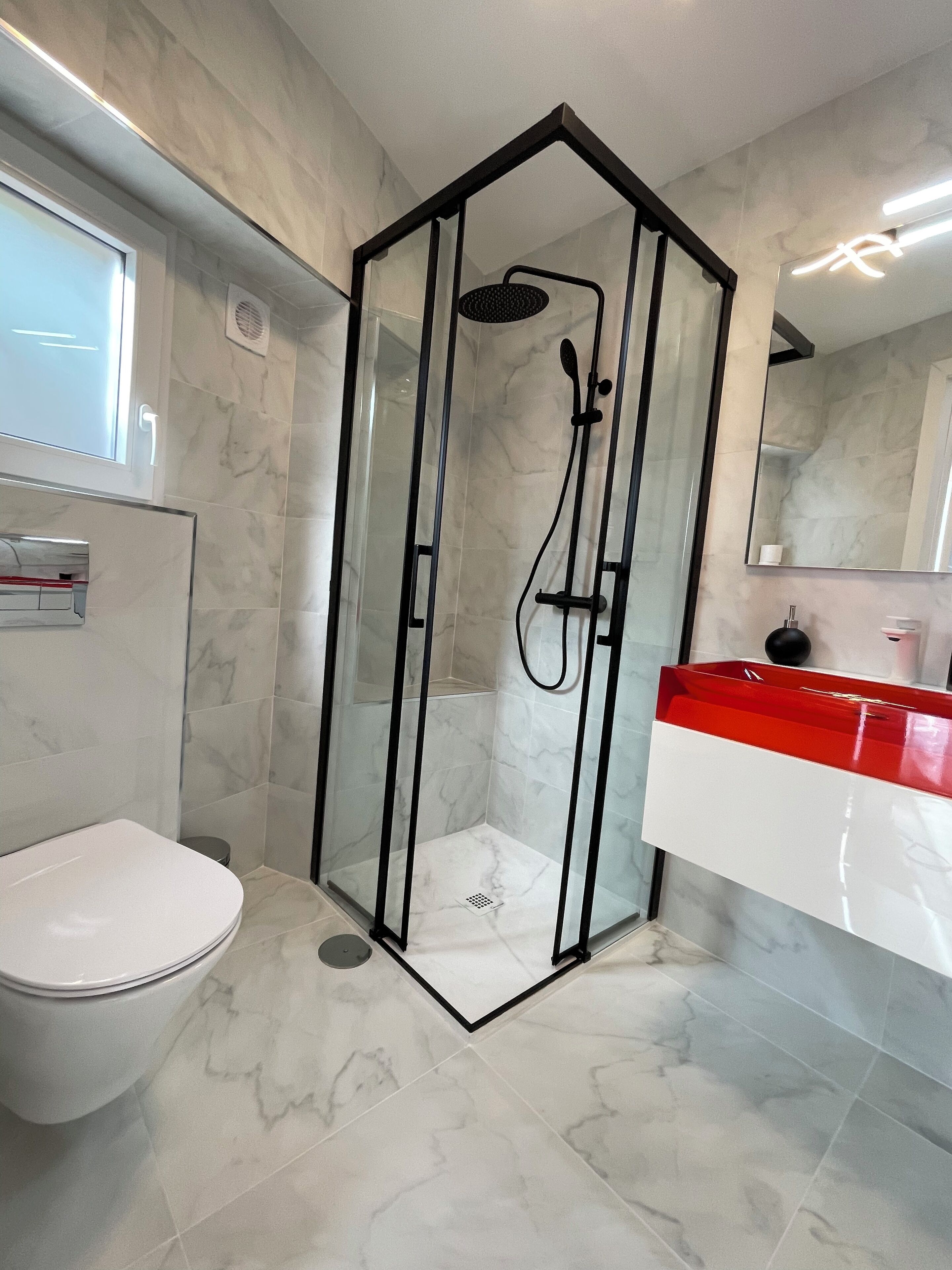 Un baño moderno con ducha envuelta en vidrio, inodoro blanco y tocador rojo y blanco.