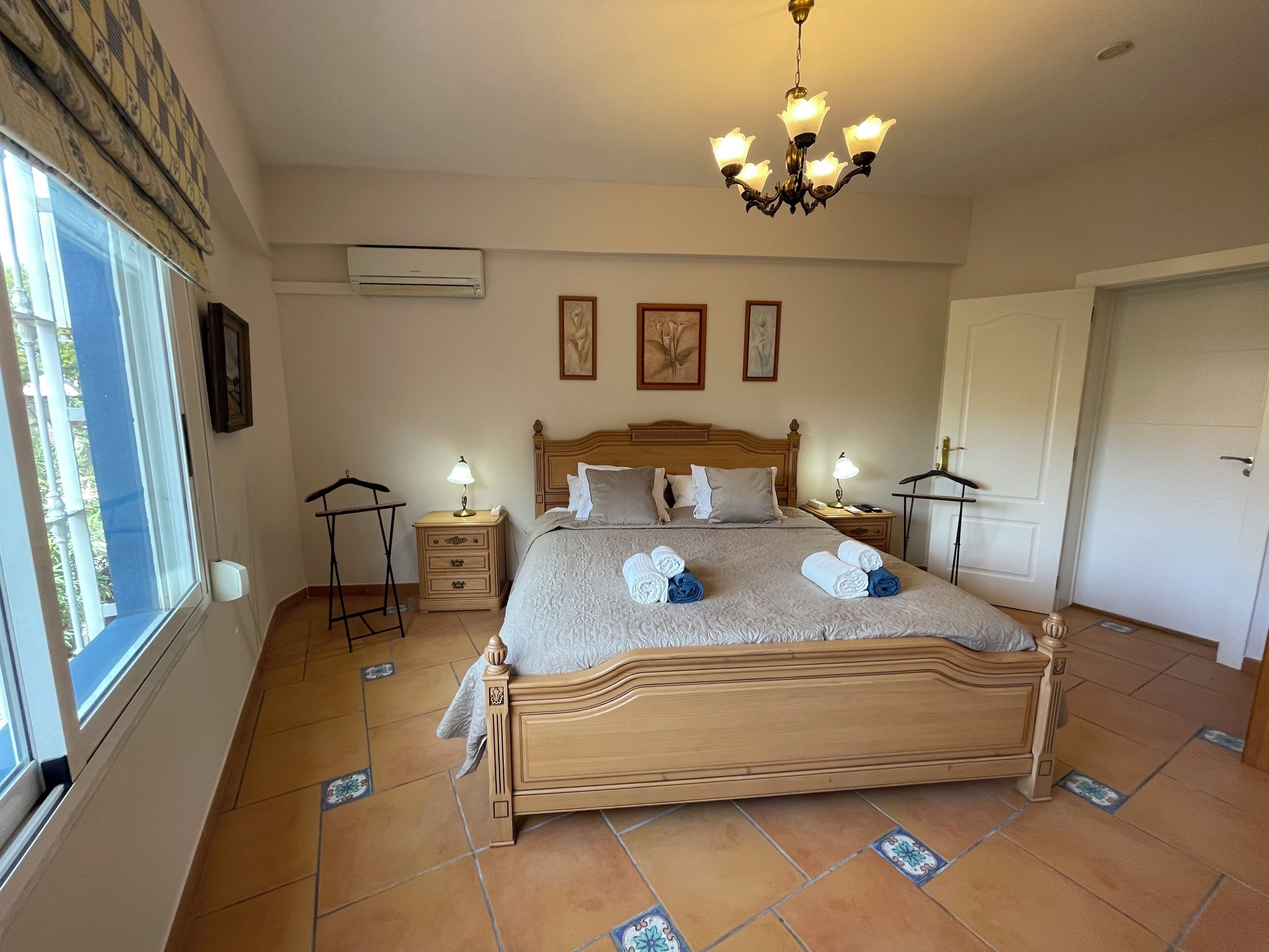 A neatly arranged bedroom with a wooden bed, bedside tables with lamps, a chandelier, and a large window.
