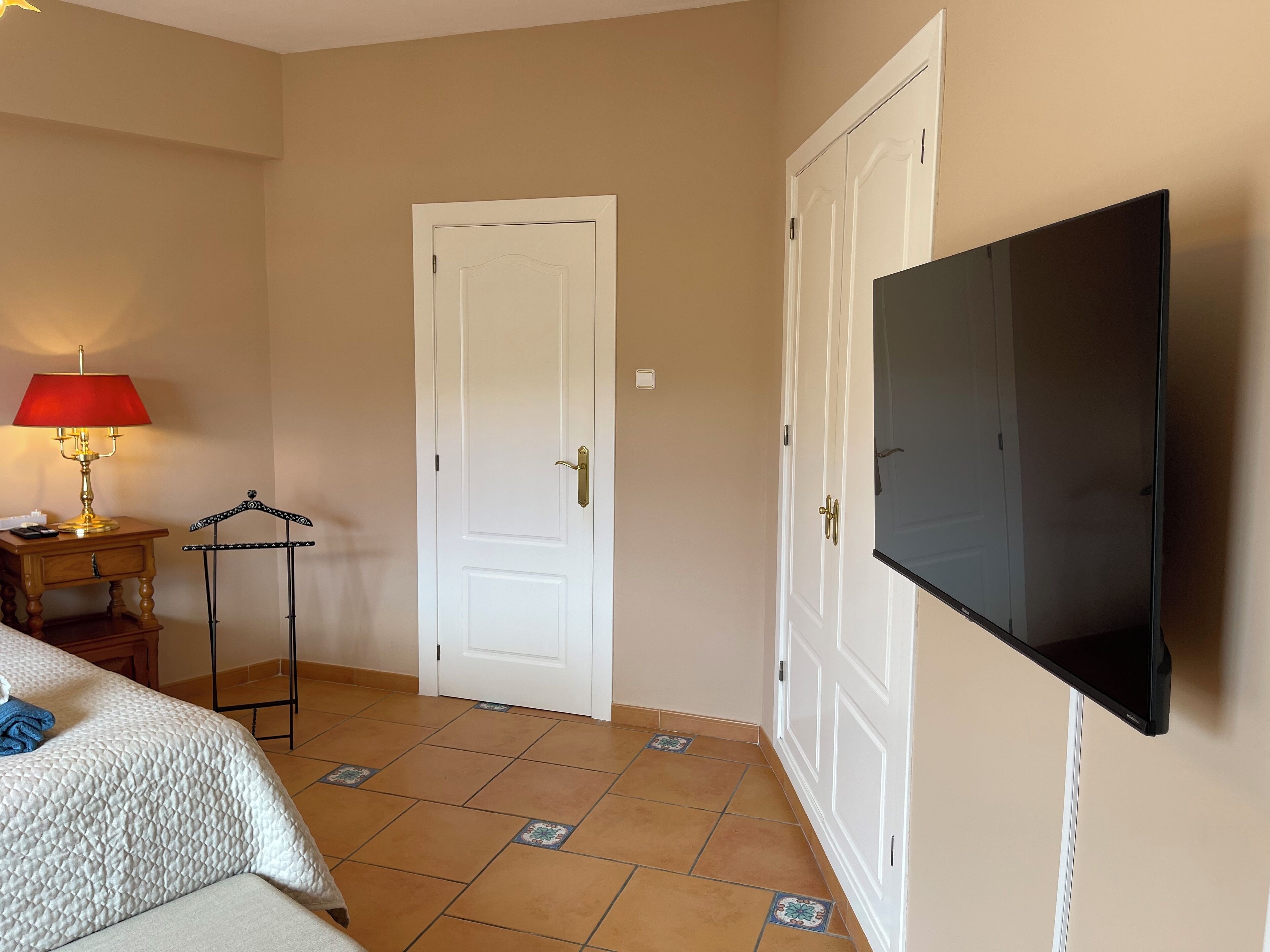 Quarto de hotel com uma cama, televisão fixada na parede, uma porta e uma mesinha com abajur.