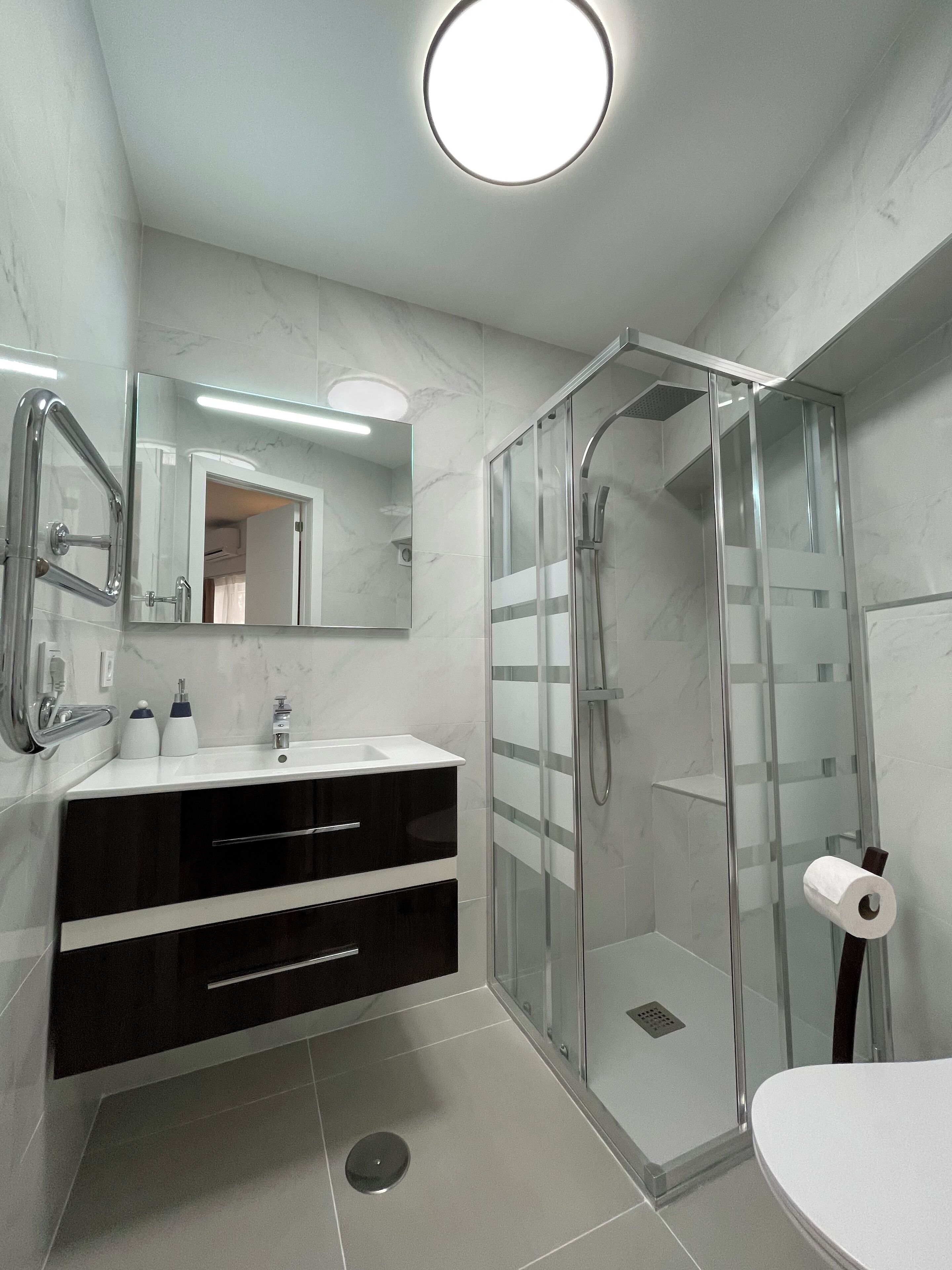 Un baño moderno con ducha envuelta en vidrio, un lavabo blanco con espejo superior y un inodoro.