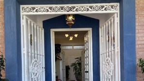 A blue-walled entrance with white decorative gates, a tiled floor, and a hallway leading to a room with a bench and potted plants. - Annas Garden (Torrevieja)