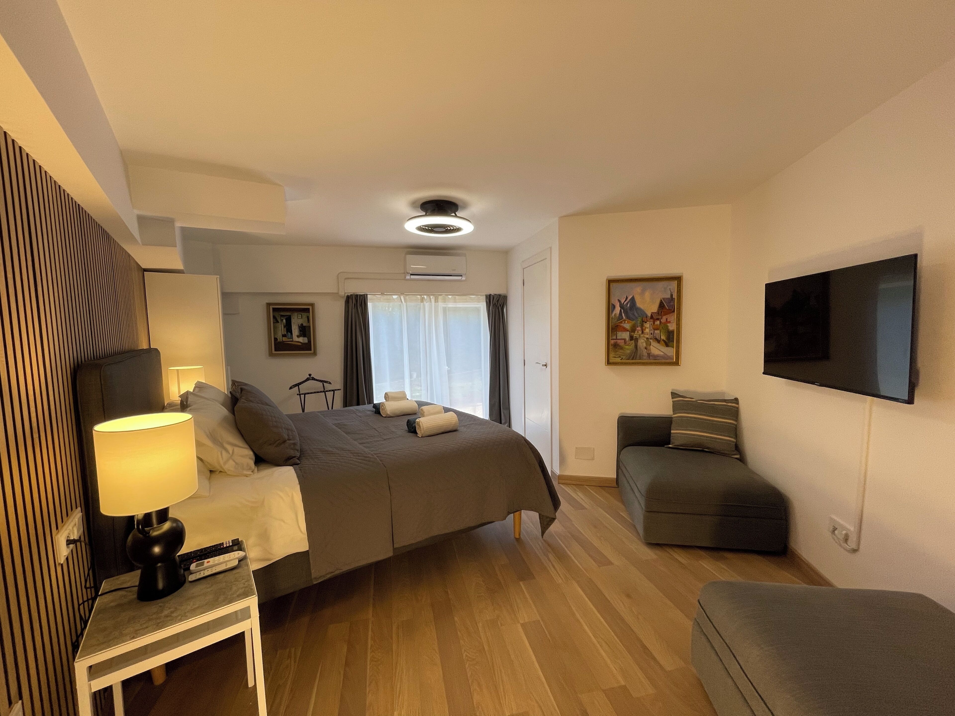 A hotel room with a bed, bedside table with a lamp, a TV, a sofa, and a painting on the wall.