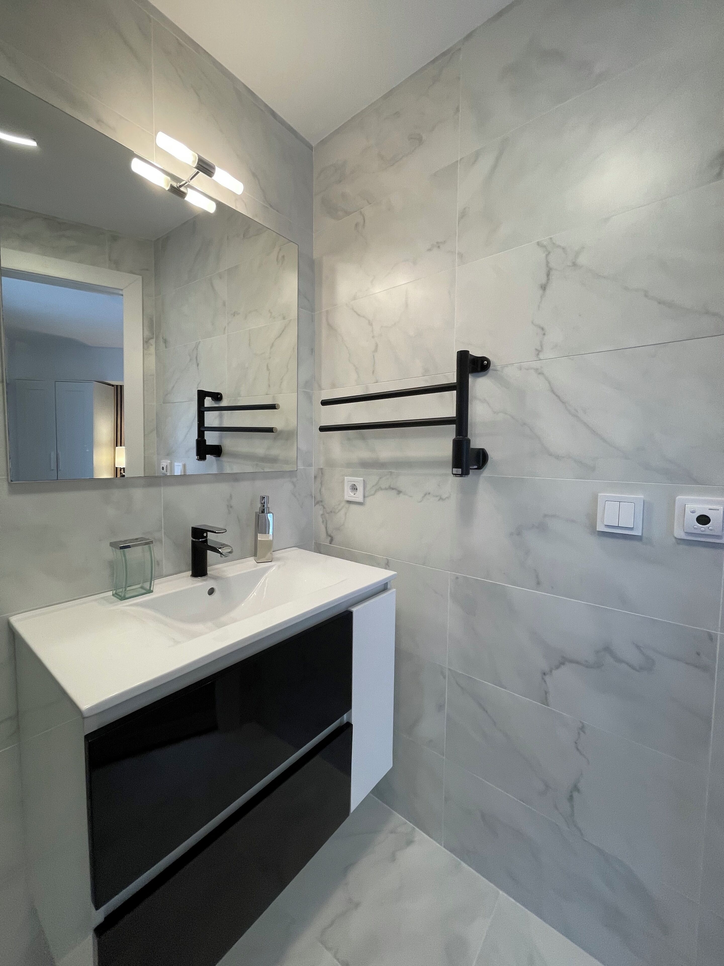 Une salle de bain moderne avec un lavabo en marbre blanc, un porte-serviettes noir et un grand miroir doté d’un éclairage intégré.