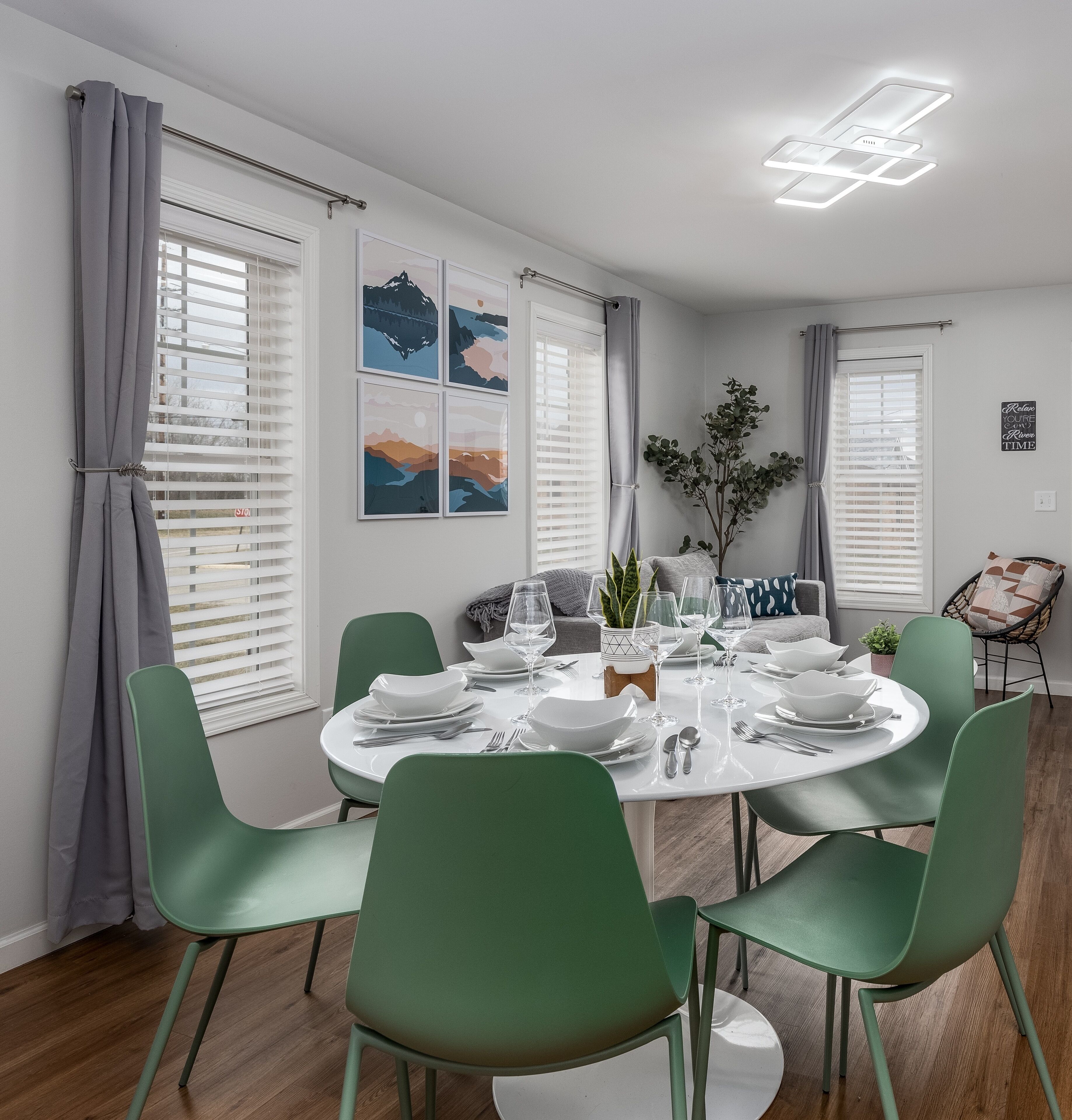 Un coin repas avec une table ronde dressée pour quatre personnes, entourée de chaises vertes. Quatre œuvres d’art encadrées ornent le mur, ainsi qu’une plante en pot et un panneau affichant une citation inspirante.