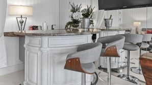 A modern kitchen with a white marble countertop, two bar stools, a flat-screen TV, and a potted plant.