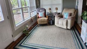 Una habitación acogedora con un ventanal grande, un sillón cómodo, una mesita con lámparas y una planta decorativa.