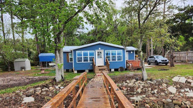 Un puente de madera que lleva a una casa azul con puerta blanca, rodeada de árboles y una cerca de madera.
