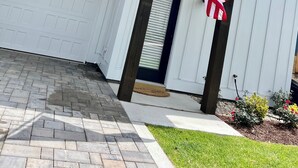 A white house with a garage door, an American flag, and a small garden with flowers. - BRAND NEW! Whalehead Beach Mediterranean Inspired Villa (Corolla)