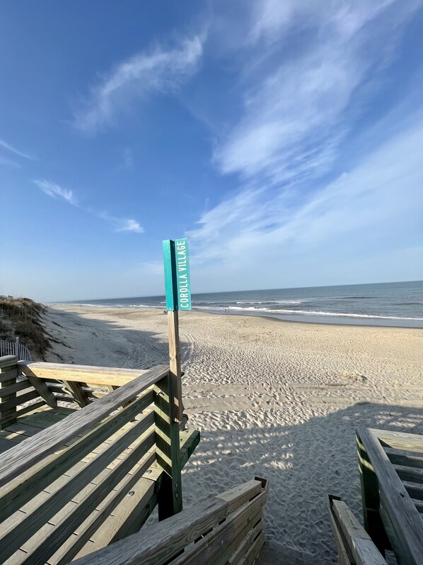 A beach with a wooden walkway leading to a sign reading "COROLLA VILLAGE".