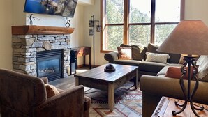 A cozy living room with a stone fireplace, a large window with a view of trees, a comfortable sofa, and a television mounted above the fireplace. - Spacious, top floor, tree views, tastefully remodeled condo, in The Village (Mammoth Lakes)