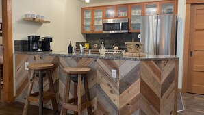 A kitchen with a herringbone-patterned backsplash, stainless steel appliances, wooden bar stools, and a coffee station.
