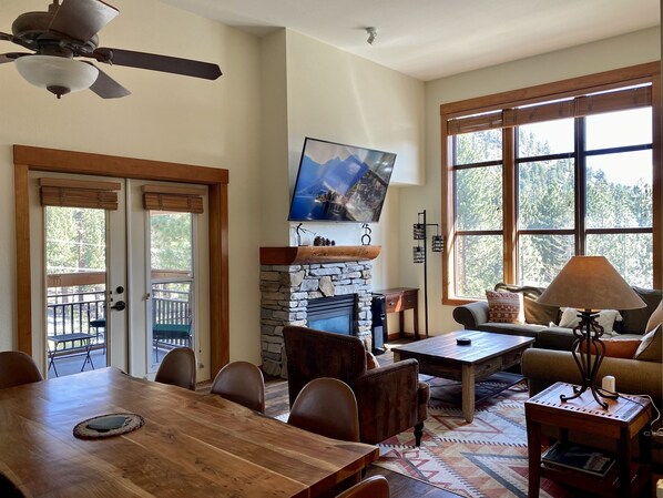 A spacious living room with a stone fireplace, a dining area, and large windows offering a view of the outdoors.