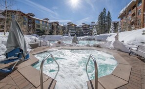 A hot tub surrounded by snow, with a pool in the background and buildings in the distance.