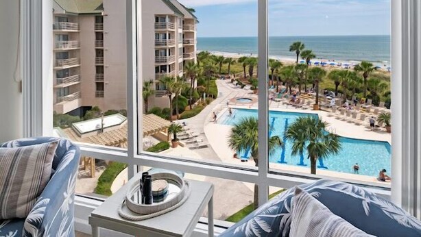 Habitación de hotel con un ventanal grande que da a una zona de piscina, una playa y un edificio con varios balcones.