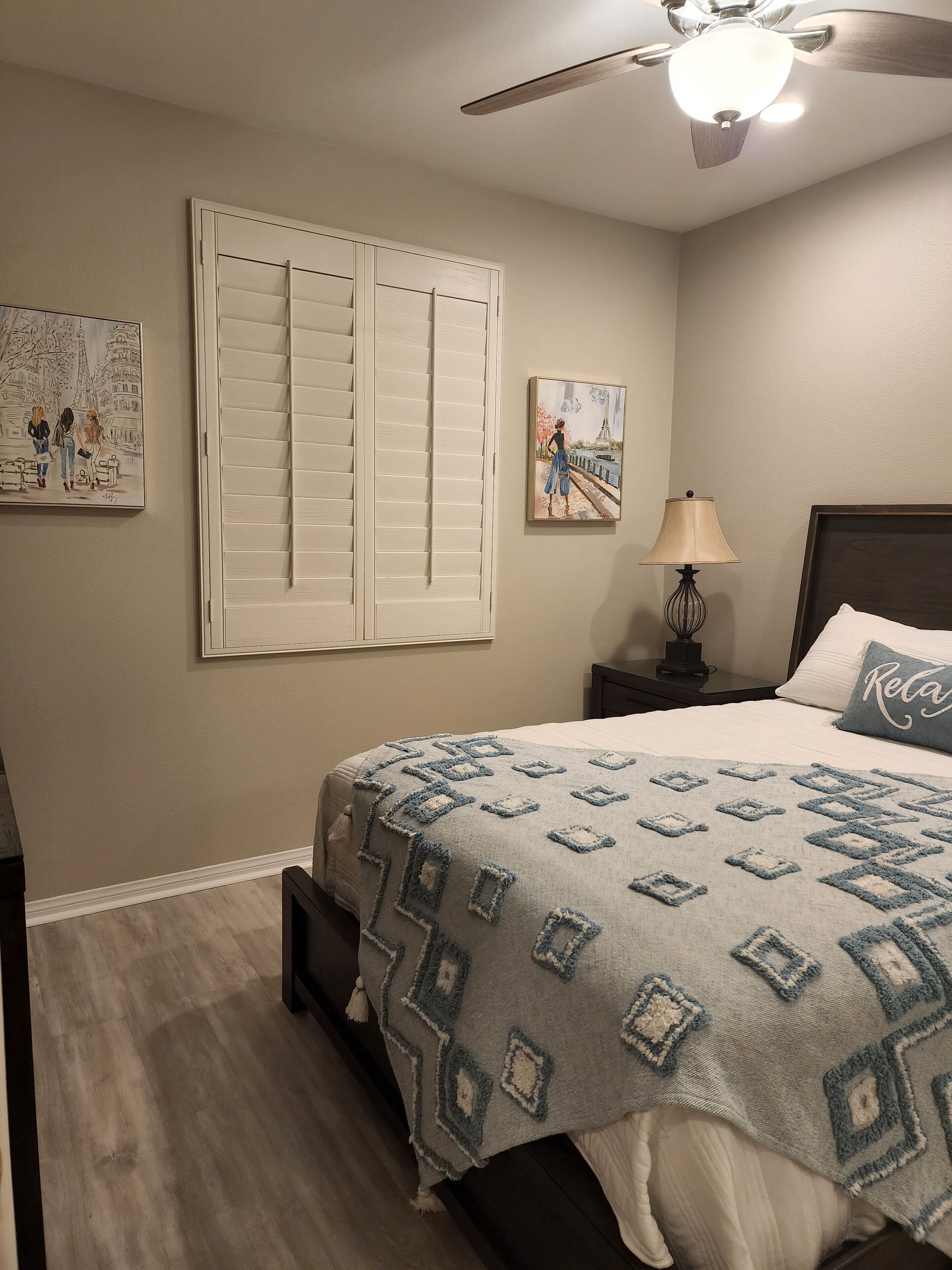 A bedroom with a bed, a nightstand, a lamp, a ceiling fan, and two paintings on the wall.