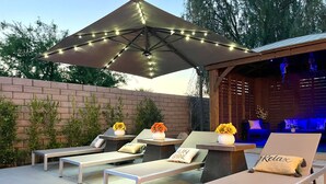 An outdoor patio with lounge chairs, a pergola with string lights, and a small seating area with cushions and a flower arrangement.