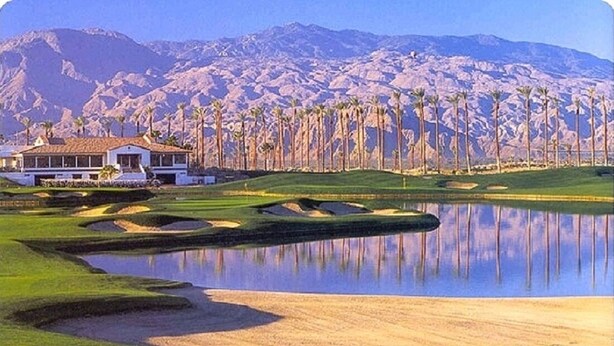 A golf course with a clubhouse, palm trees, and mountains in the background.