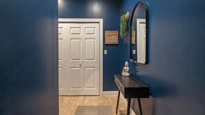 Um corredor com uma porta branca, um aparador preto com espelho e uma placa de boas-vindas na parede.