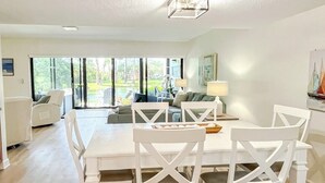 Un comedor amplio con una mesa y sillas blancas, una lámpara en el techo y vistas al exterior a través de grandes ventanales.