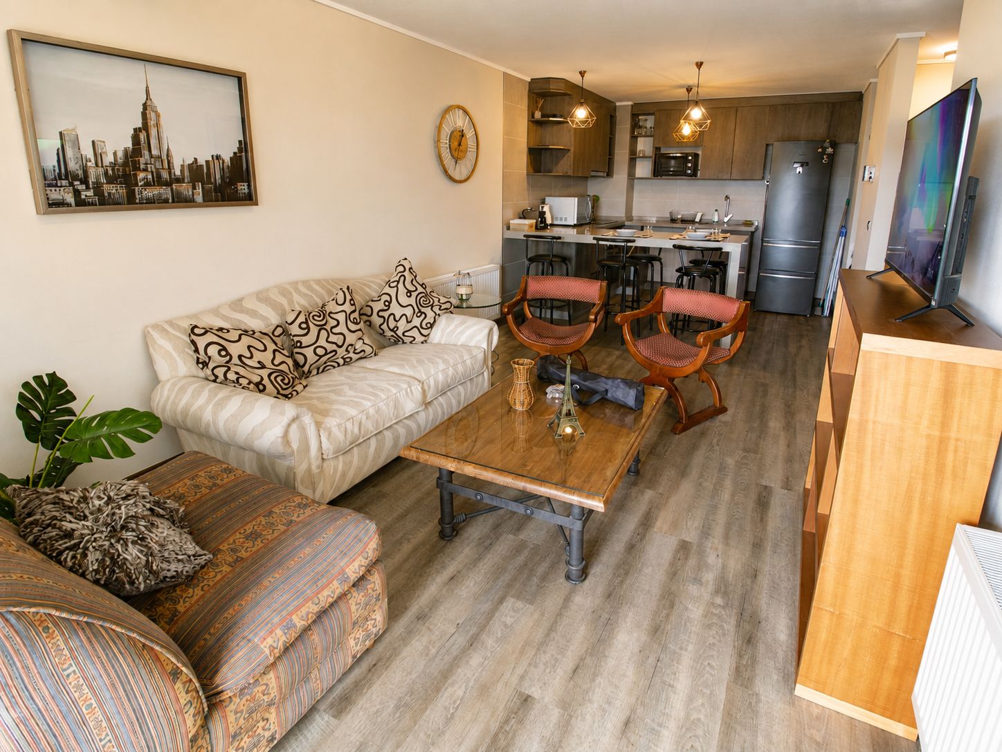 A living room with a sofa, two armchairs, a coffee table, and a dining table. There is a kitchen in the background with a refrigerator, microwave, and cabinets.