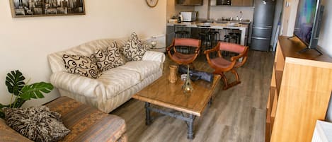 A living room with a sofa, two armchairs, a coffee table, and a dining table. There is a kitchen in the background with a refrigerator, microwave, and cabinets.