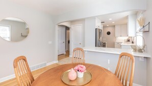 Un comedor con una mesa de madera, cuatro sillas, un espejo redondo y una cocina al fondo.