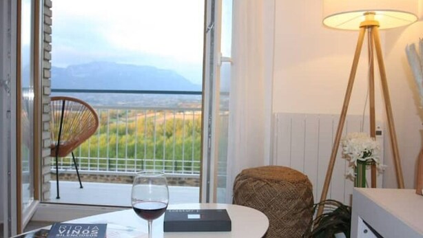 Una habitación con vistas a las montañas, una mesa con una copa de vino tinto, una silla y una lámpara.