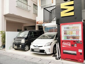 A parking area with two cars, a red Coca-Cola vending machine, and a building with balconies.