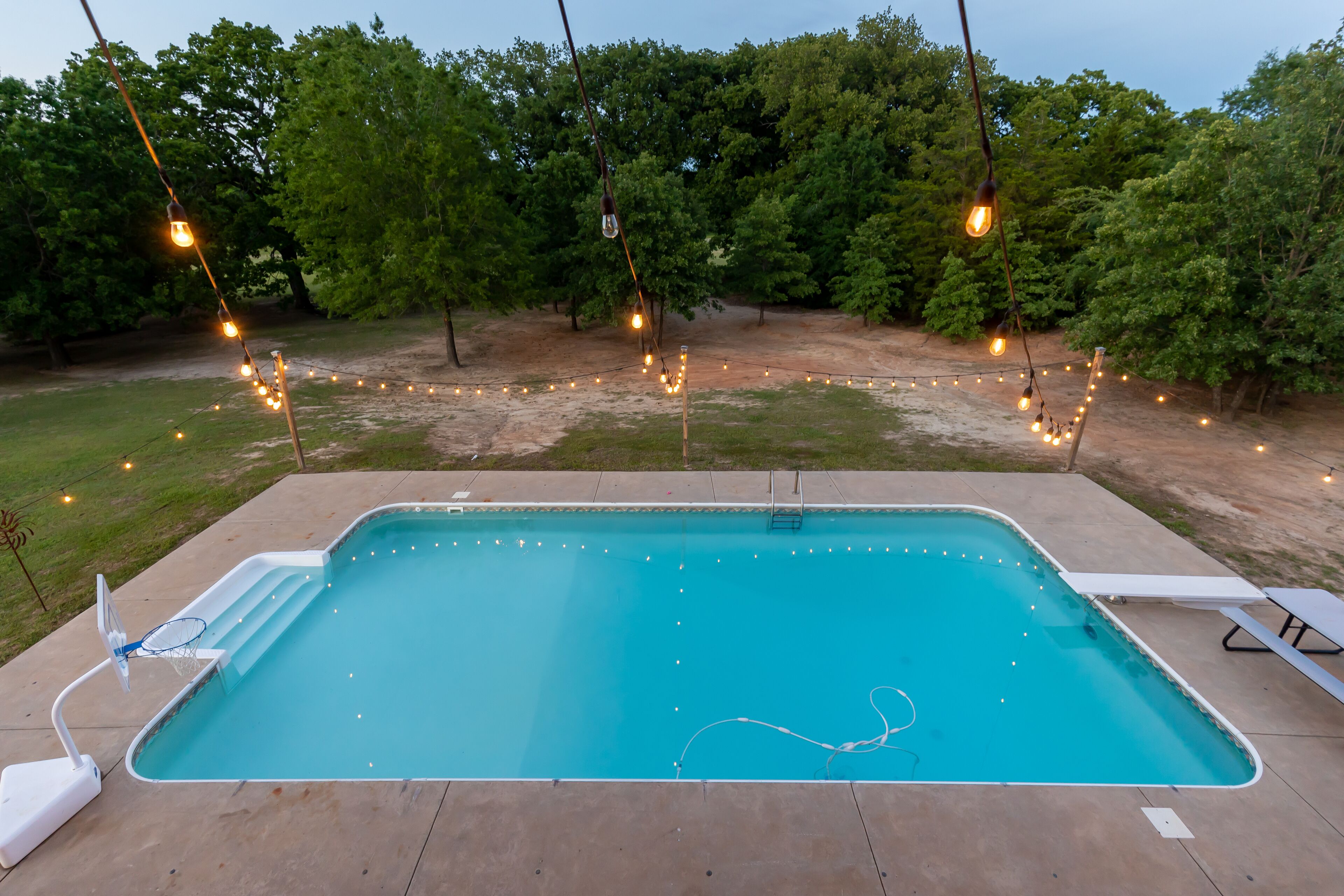 En rektangulär swimmingpool med en stege på ena sidan, omgiven av en betongplattform. På plattformen finns solstolar och ett picknickbord.