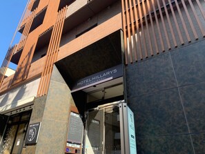 Entrada de hotel con fachada moderna, un cartel que dice 'Hotel Hillaries' y un ascensor visible.