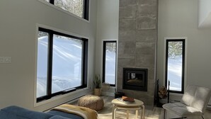A modern living room with a large blue sofa, a fireplace, and large windows offering a view of the outdoors. - La Cache à Théo (Lac-Supérieur)