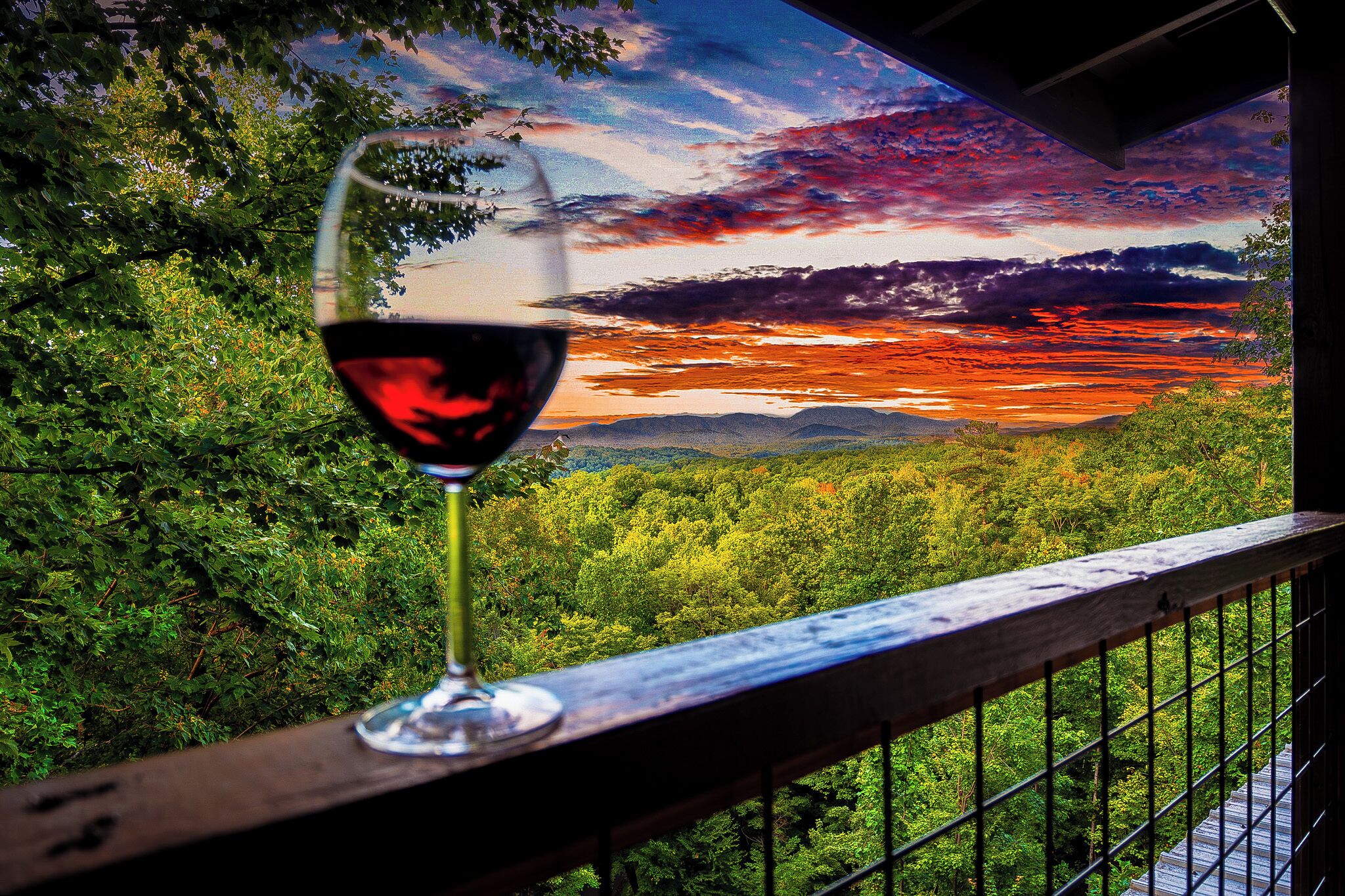 A glass of red wine on a balcony with a scenic view of a forest and a sunset.
