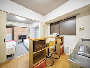 Quarto de hotel compacto com cama, pequena área de cozinha, mesa de madeira com cadeiras e televisão.
