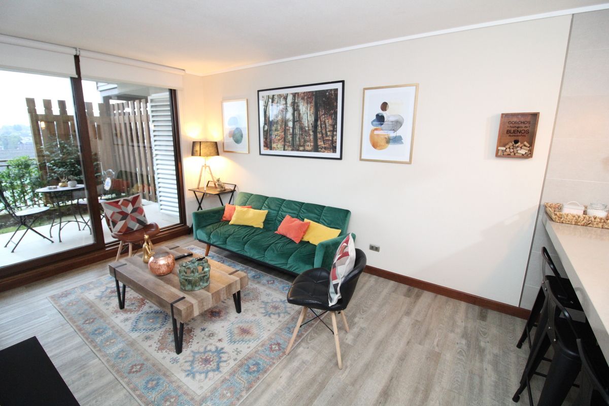 A living room with a green sofa, a coffee table, and a chair. There are framed pictures on the wall and a window with a view.
