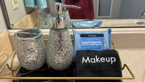 Un tocador de baño con encimera de mármol negro, un atril para maquillaje con marco dorado, dos jarrones de vidrio decorativos, una bolsa para removedor de maquillaje y una toalla con la palabra 'Maquillaje'.