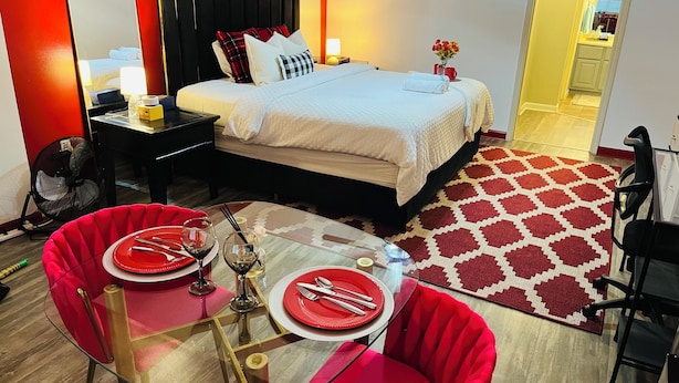 Habitación de hotel con cama, mesita de noche, lámpara y una zona de comedor con mesa de vidrio y sillas rojas.