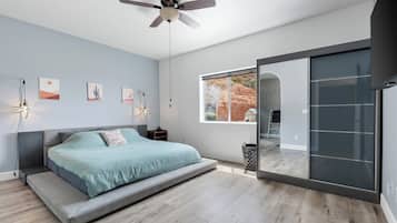 A modern bedroom with a bed, ceiling fan, artwork, and a large window.