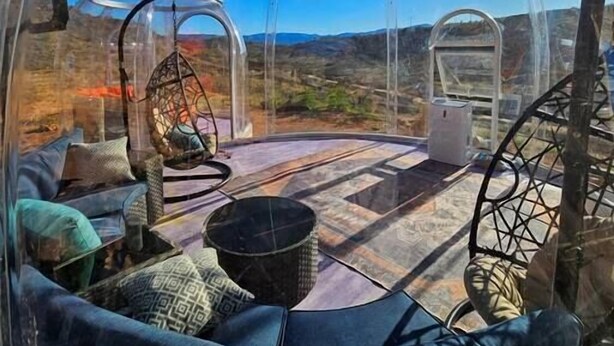 Una estructura tipo cúpula transparente con un ventanal circular, una silla colgante y un sofá con cojines estampados.