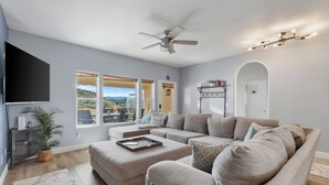 Una sala de estar moderna con un sofá modular, un televisor de pantalla plana, un ventilador de techo y ventanas amplias que ofrecen vistas al exterior.
