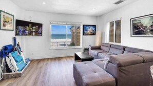 A living room with a large window offering a view of the beach, a brown leather sofa, and a wheelchair in the corner. - Beachfront 1BR · Ocean View, Pool + Free Parking (Hollywood)