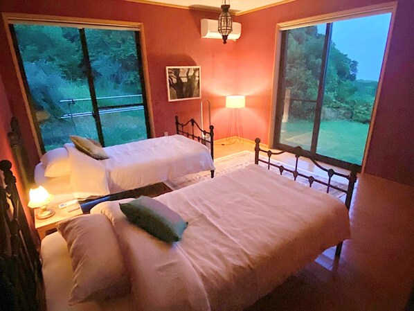 Ein Schlafzimmer mit zwei Einzelbetten, einem Nachttisch mit Lampe und großen Fenstern mit Blick ins Freie.