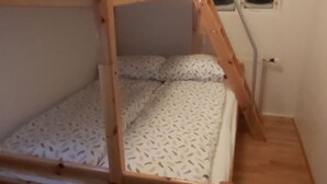 Een stapelbed met houten frame en een matras met bladpatroon. Er is een raam met erboven een groen vluchtlicht, en een ladder die naar het bovenste bed leidt.