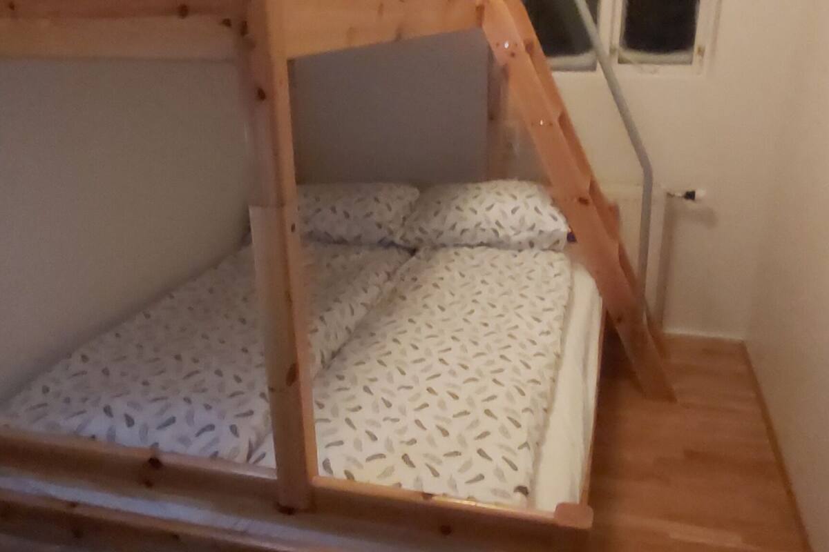 Een stapelbed met houten frame en een matras met bladpatroon. Er is een raam met erboven een groen vluchtlicht, en een ladder die naar het bovenste bed leidt.