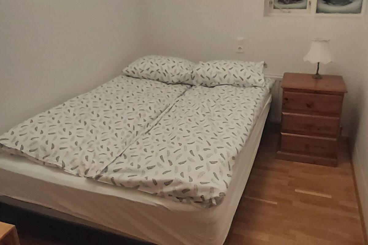 Een slaapkamer met een bed, houten vloer, nachtkastje met een lamp en een raam met een groen bord waarop nooduitgang staat.