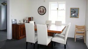 Een eetgedeelte met een houten tafel en witte stoelen, een klein kastje en een schilderij aan de muur.