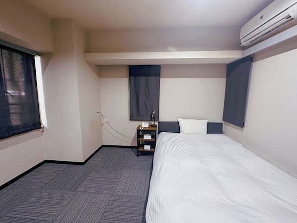 A hotel room with a bed, a bedside table with a lamp, a telephone, and an air conditioning unit.